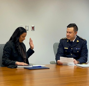 Cpl. Dhesi sitting at a table with a male officer. Her left hand is raised.