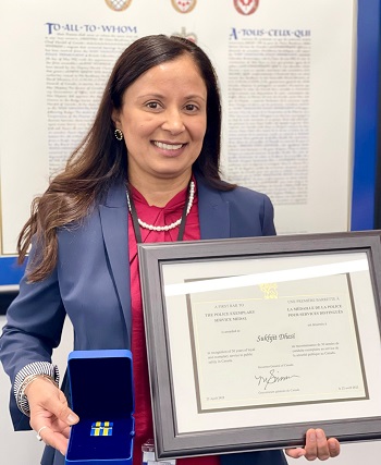 Cpl. Dhesi wearing a blue blazer and a red shirt, holding a framed award and a small velvet box. 